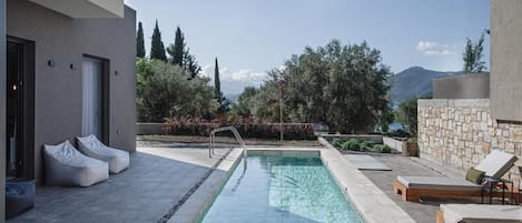 A swimming pool with a stone wall and lounge chairs, surrounded by vegetation and a mountainous backdrop.