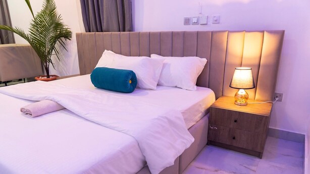 Een net opgemaakt bed met witte lakens en een blauw sierkussen, een bedlamp en een klein houten nachtkastje.