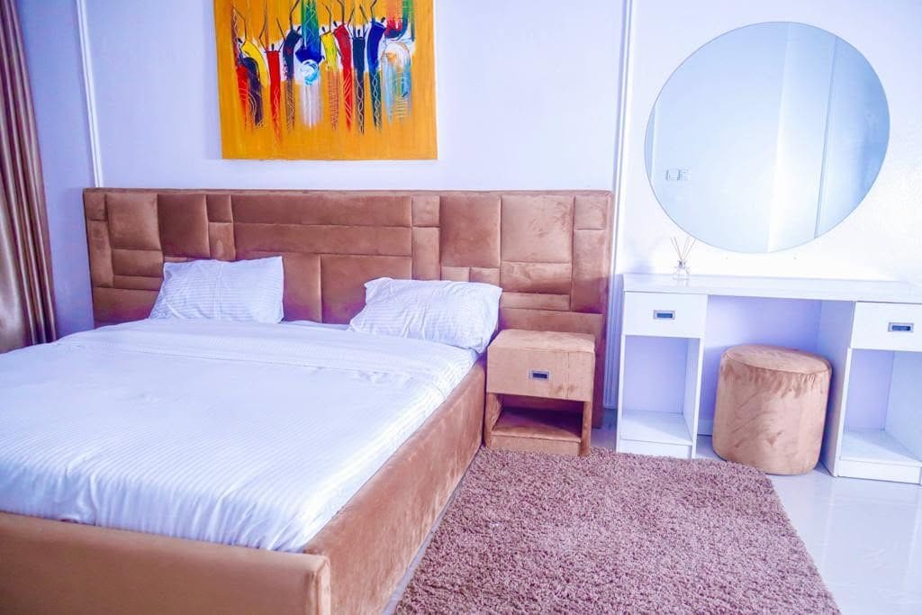 Un dormitorio con una cama grande, una mesita de noche de madera, un espejo redondo y un cuadro abstracto y colorido en la pared.