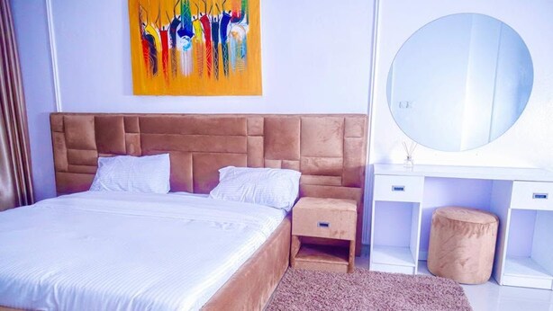 A bedroom with a large bed, a wooden nightstand, a round mirror, and a colorful abstract painting on the wall. - Ken Kol Apartment  (Lekki)