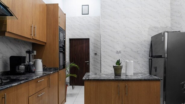 A compact kitchen with wooden cabinets, a built-in oven, a refrigerator, and a countertop with a coffee maker and a potted plant. - Ken Kol Apartment  (Lekki)