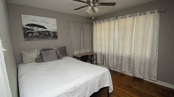 Un dormitorio con cama, escritorio, ventilador de techo y ventana con cortinas.