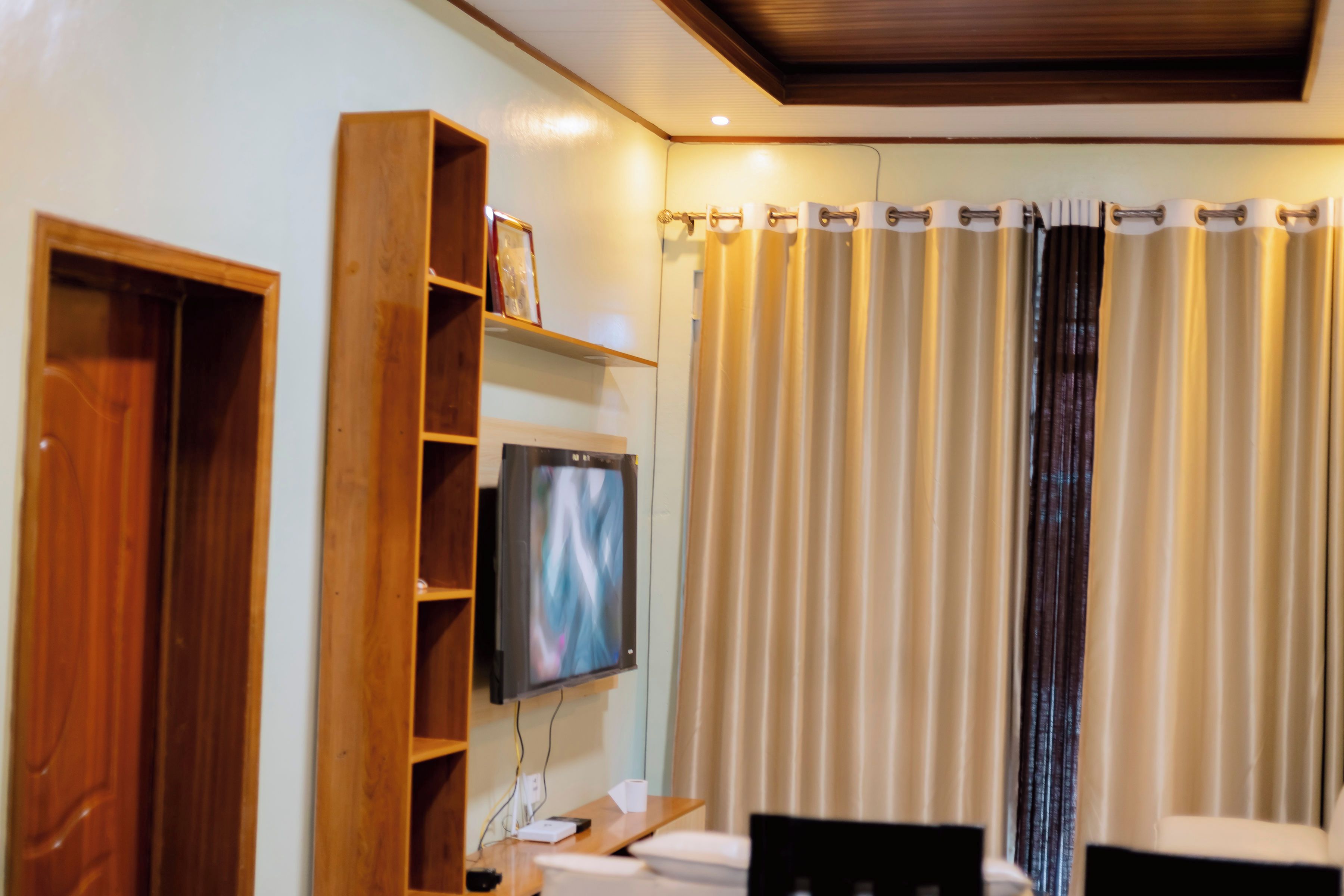 A hotel room with a wooden wardrobe, a television mounted on the wall, a desk with a chair, and a curtain with a patterned design.
