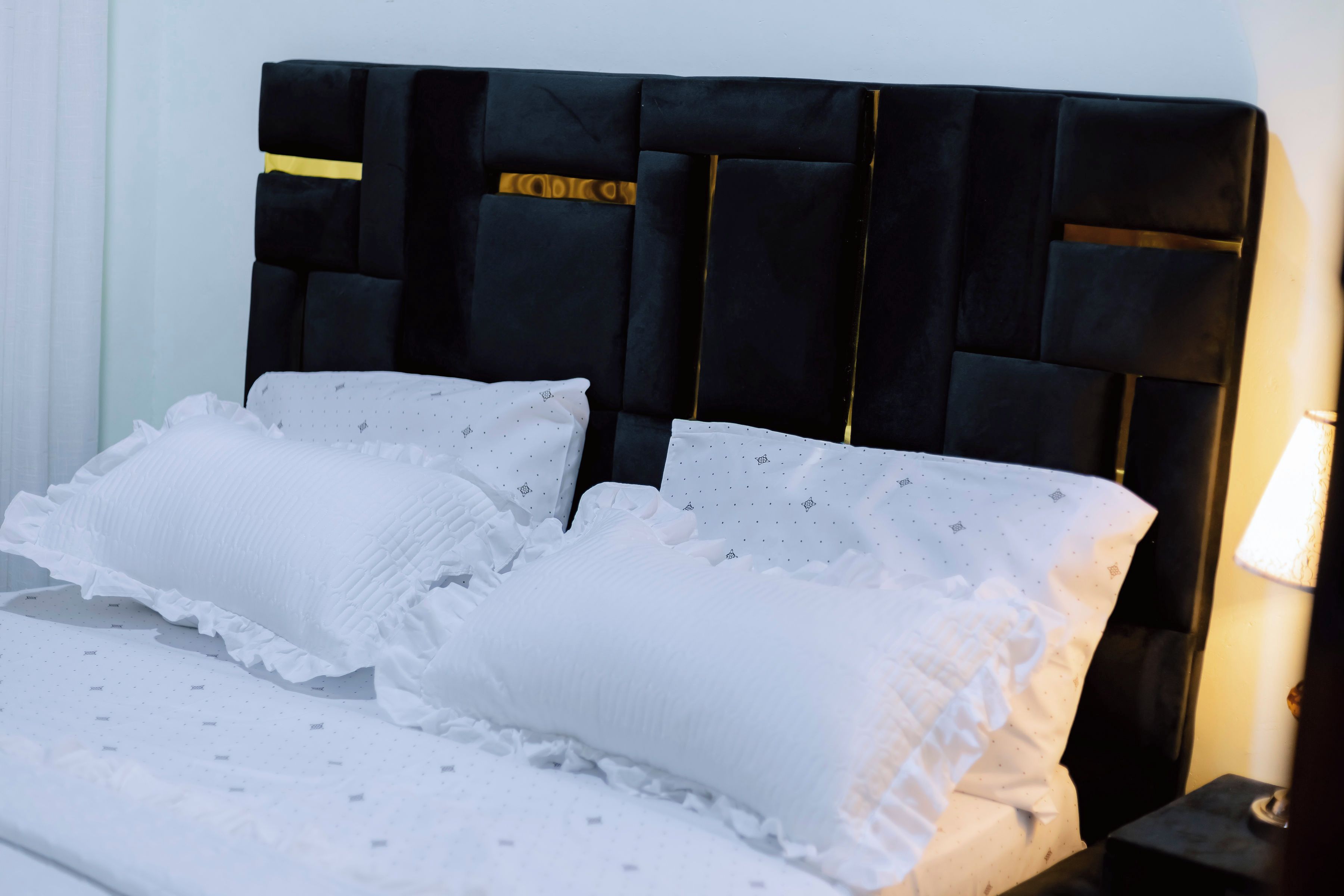 Una cama con cabecera tapizada de color negro, almohadas blancas y una colcha blanca con motivos pequeños.