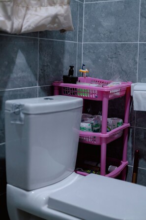 Un baño con inodoro blanco, una cesta rosa con artículos de tocador y una toalla colgada en la pared.