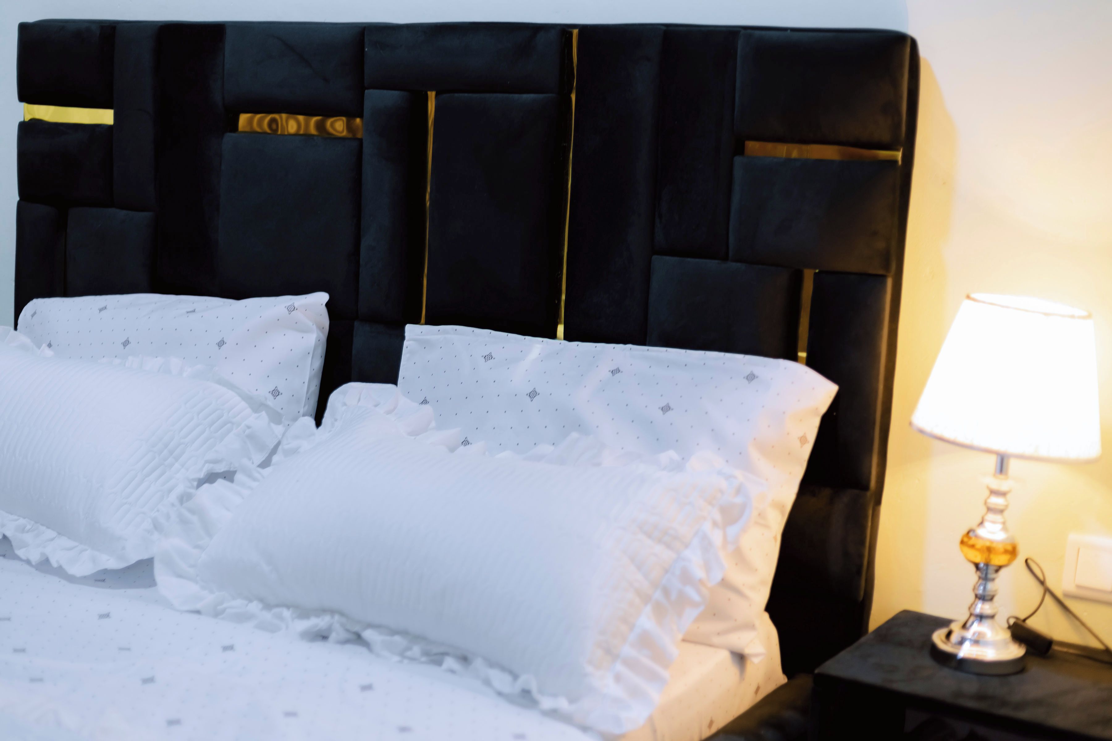 A bed with a black upholstered headboard, white pillows with ruffled edges, and a bedside lamp on a nightstand.