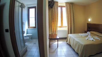 A hotel room with a bed, a bedside table, a window with curtains, a bathroom with a toilet and sink, and a door leading to another room.