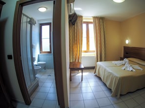 Habitació d'hotel amb llit, taula de nit, finestra amb cortines, bany amb vàter i rentapans, i porta que dona a una altra habitació.