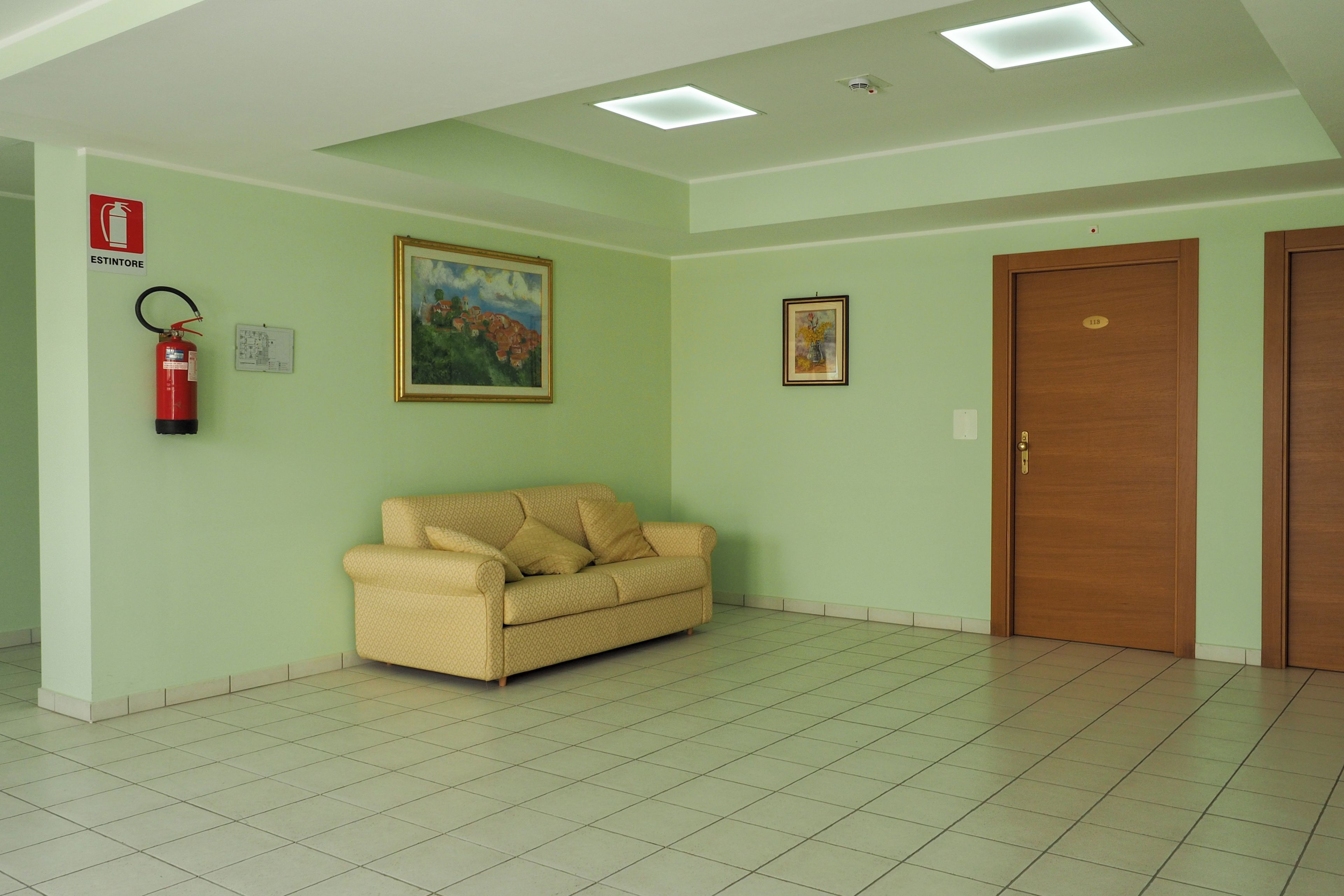 A room with a beige sofa, a painting, a fire extinguisher, and a door with a number plate.