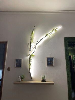 Eine Wandgestell-Verzweigung mit Blättern, beleuchtet von einer Leuchte, ein kleiner Topfpflanze und zwei gerahmte Bilder.