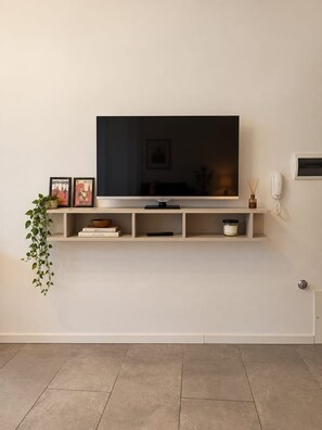 A flat-screen TV mounted on a wall with a shelf below containing books, a remote, and a small decorative item. - The Court of the Big Fish (Arezzo)