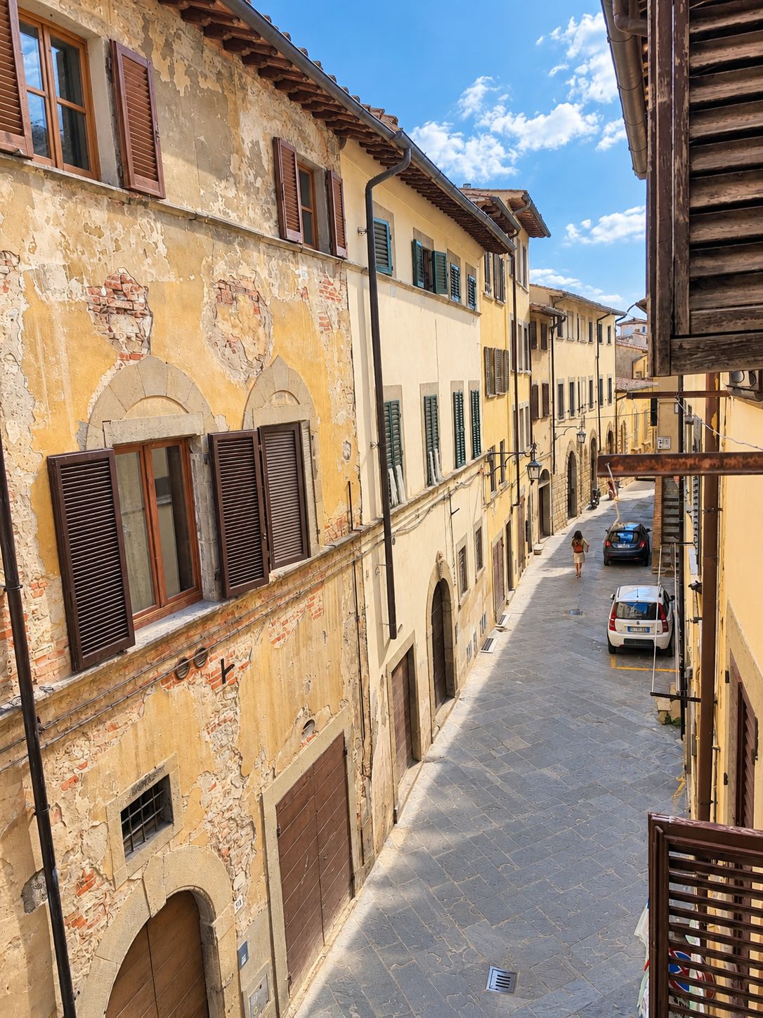 A narrow street with buildings on either side, featuring arched doorways and shuttered windows.