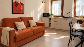 A living room with a sofa, a dining table with chairs, a kitchen area with a microwave, and a poster on the wall.