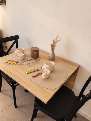 Ein Holztisch mit einer beigen Tischmatte, zwei weißen Tassen, einer braunen Kanne, einer Vase mit Trockenblumen und zwei Tüten mit Snacks.