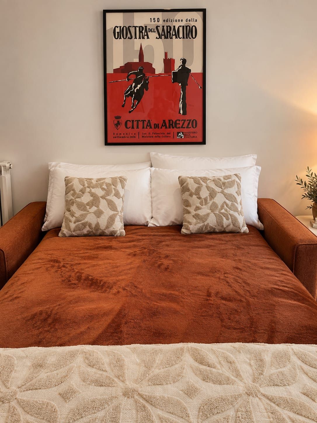 A bed with a brown cover and two patterned pillows, a framed poster on the wall, and a bedside plant.