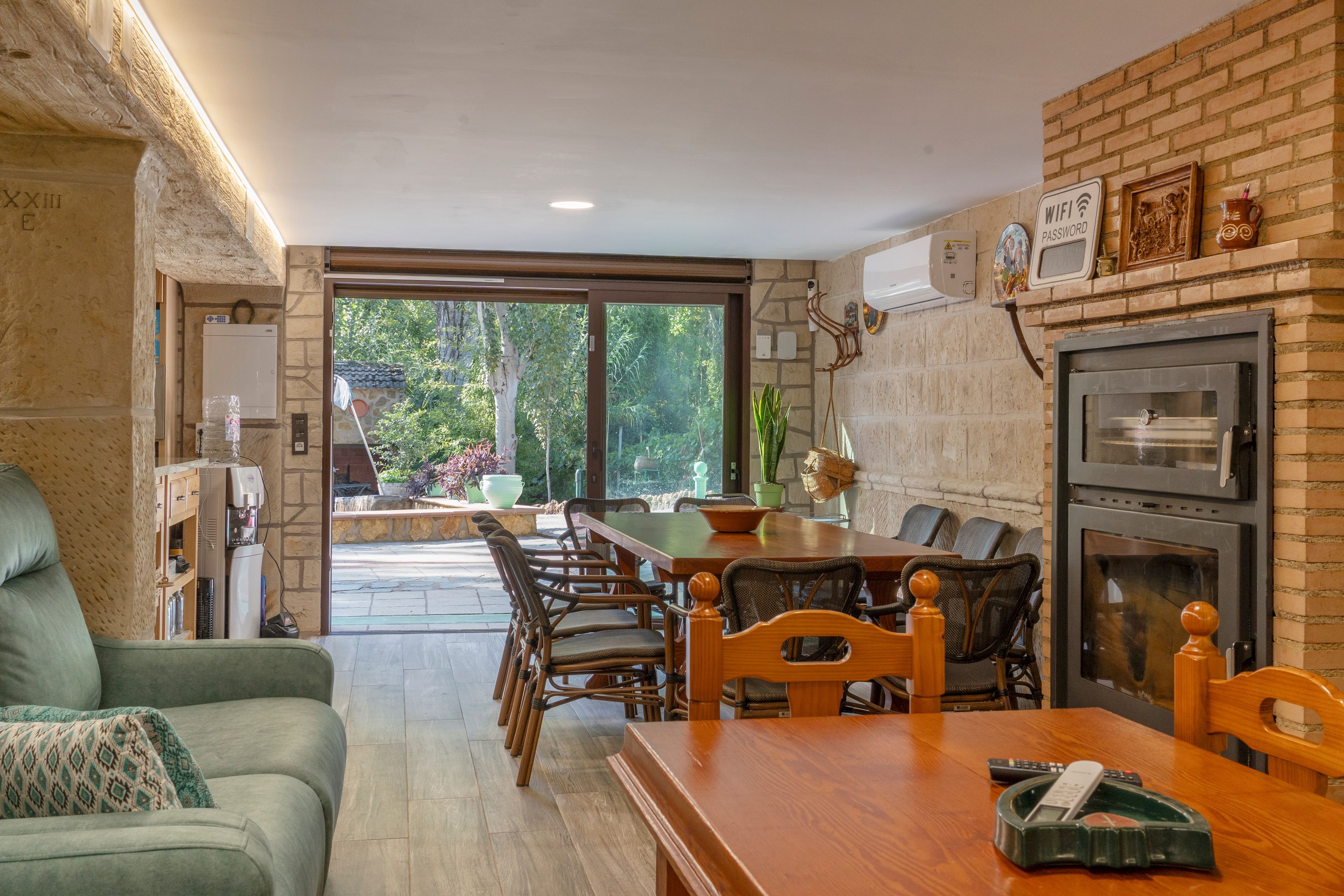 Un comedor con mesa y sillas de madera, pared de ladrillo y vista a un espacio exterior por un ventanal.
