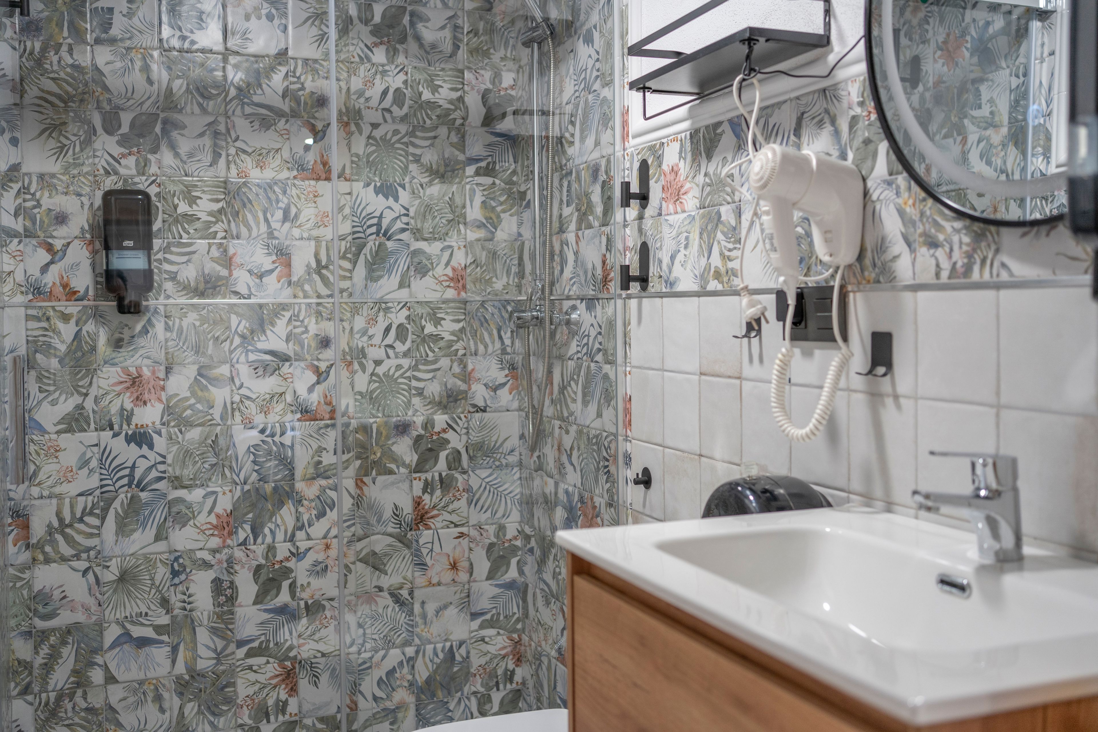 Un baño con baldosas de estampado floral, un lavabo blanco, un cabezal de ducha de mano y un secador de pelo montado en la pared.