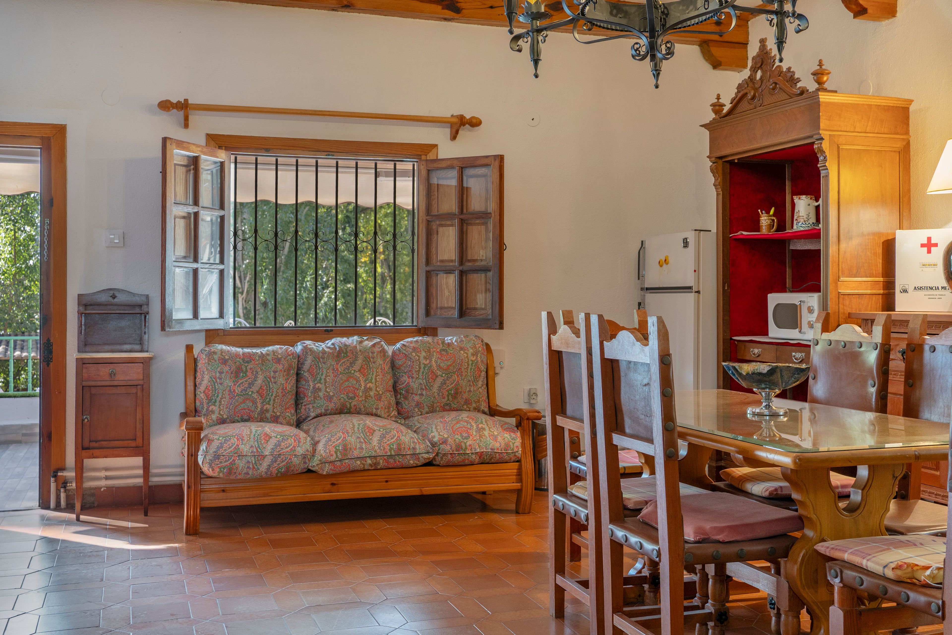 Un salon chaleureux avec un canapé en bois, une table à manger avec des chaises, un lustre et une fenêtre donnant sur un paysage verdoyant.