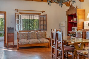 A cozy living room with a wooden sofa, a dining table with chairs, a chandelier, and a window with a view of greenery.