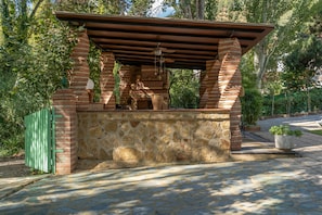 A stone and brick outdoor structure with a roof, possibly a barbecue area, surrounded by greenery.