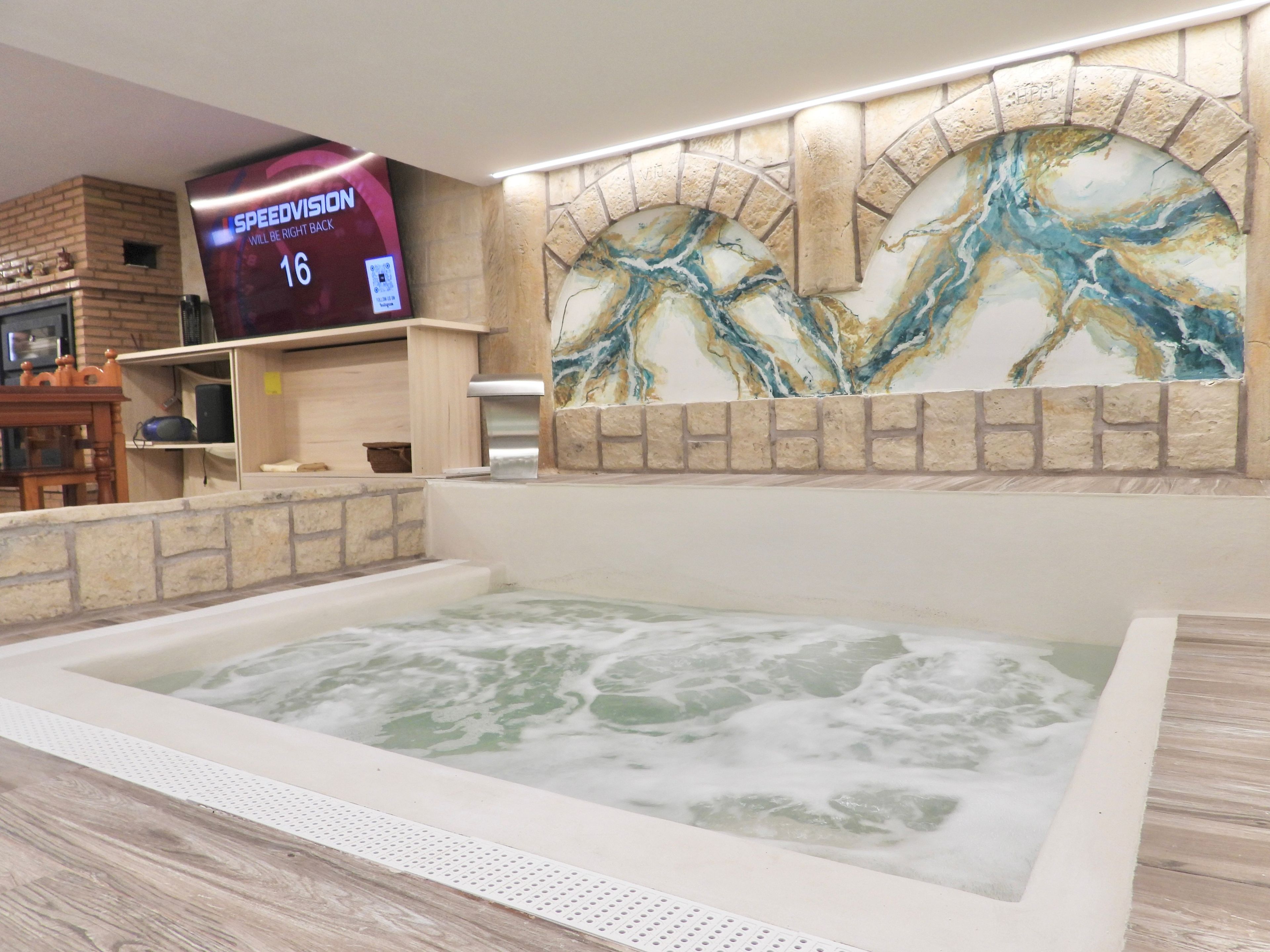 Un área de spa con un jacuzzi, un televisor mostrando 'SPEEDVISION', y una pared decorativa con un diseño similar al mármol.