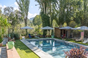 A swimming pool with lounge chairs and umbrellas, surrounded by greenery and stone walls.