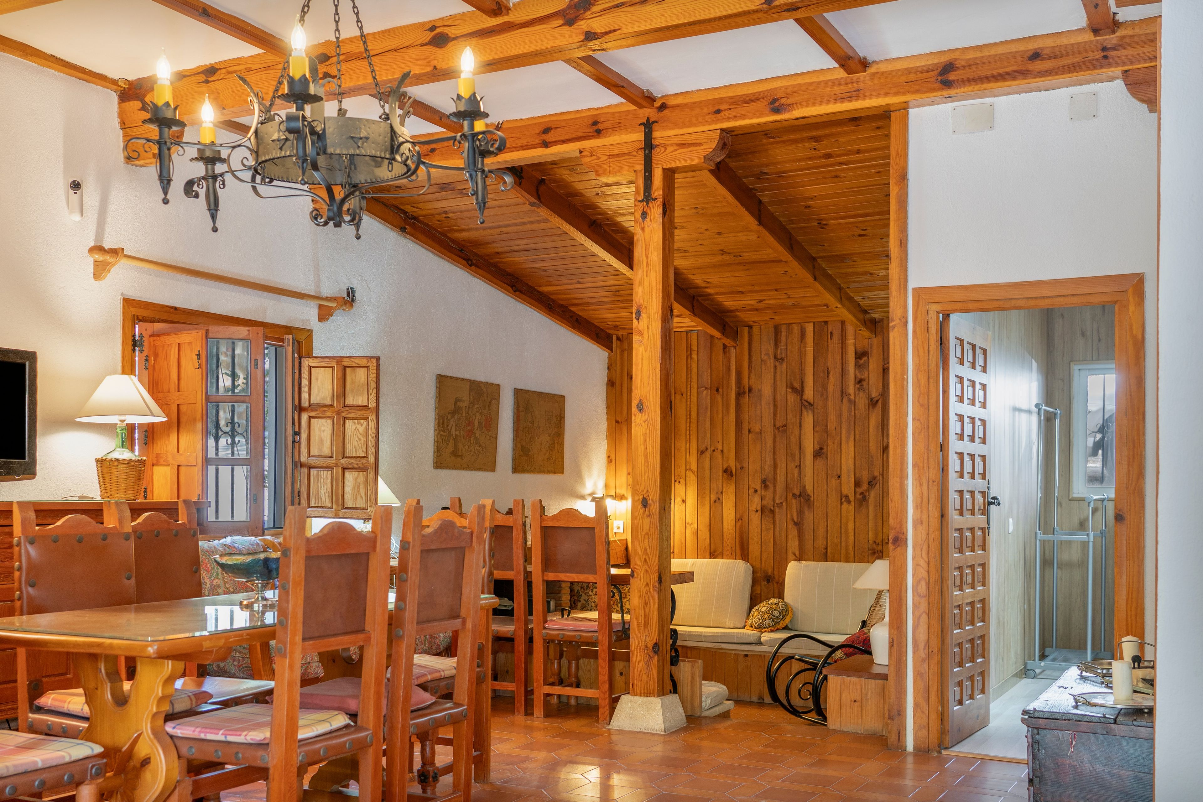 Un interior rústico con vigas de madera, una araña, un comedor con sillas y mesa de madera, y un sofá.