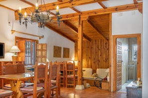 A rustic interior with wooden beams, a chandelier, a dining area with wooden chairs and a table, and a sofa.