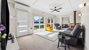 Ein modernes Wohnzimmer mit einem grauen Sofa, einem gelben Sessel und einem kleinen Tisch mit einer Pflanze. Zwei Türen führen zu einem Balkon, und ein Deckenventilator ist sichtbar.