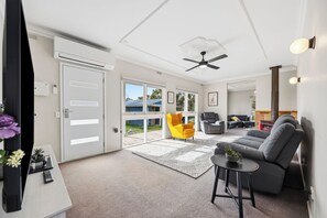 A modern living room with a grey sofa, a yellow armchair, and a small table with a plant. There are two doors leading to a balcony, and a ceiling fan is visible. - Spacious 5-bedroom 6 acres country house  with AC & dogs run ( pet friendly) (Stawell)