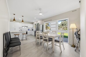 A modern kitchen with white cabinets, a dining area with a table and chairs, and a view of the outdoors through large windows. - Spacious 5-bedroom 6 acres country house  with AC & dogs run ( pet friendly) (Stawell)
