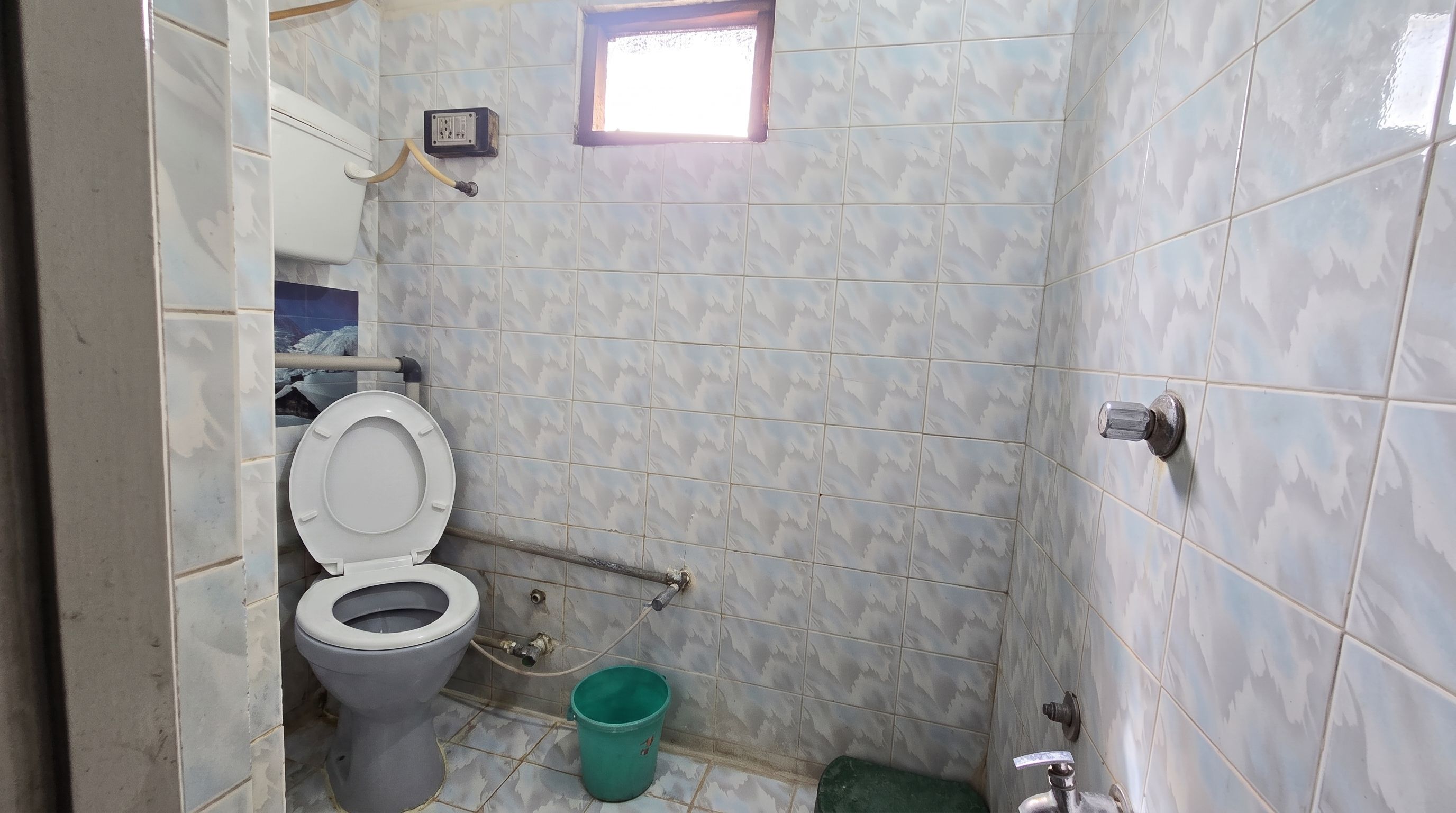 A small bathroom with a toilet, a green bucket, and a window.