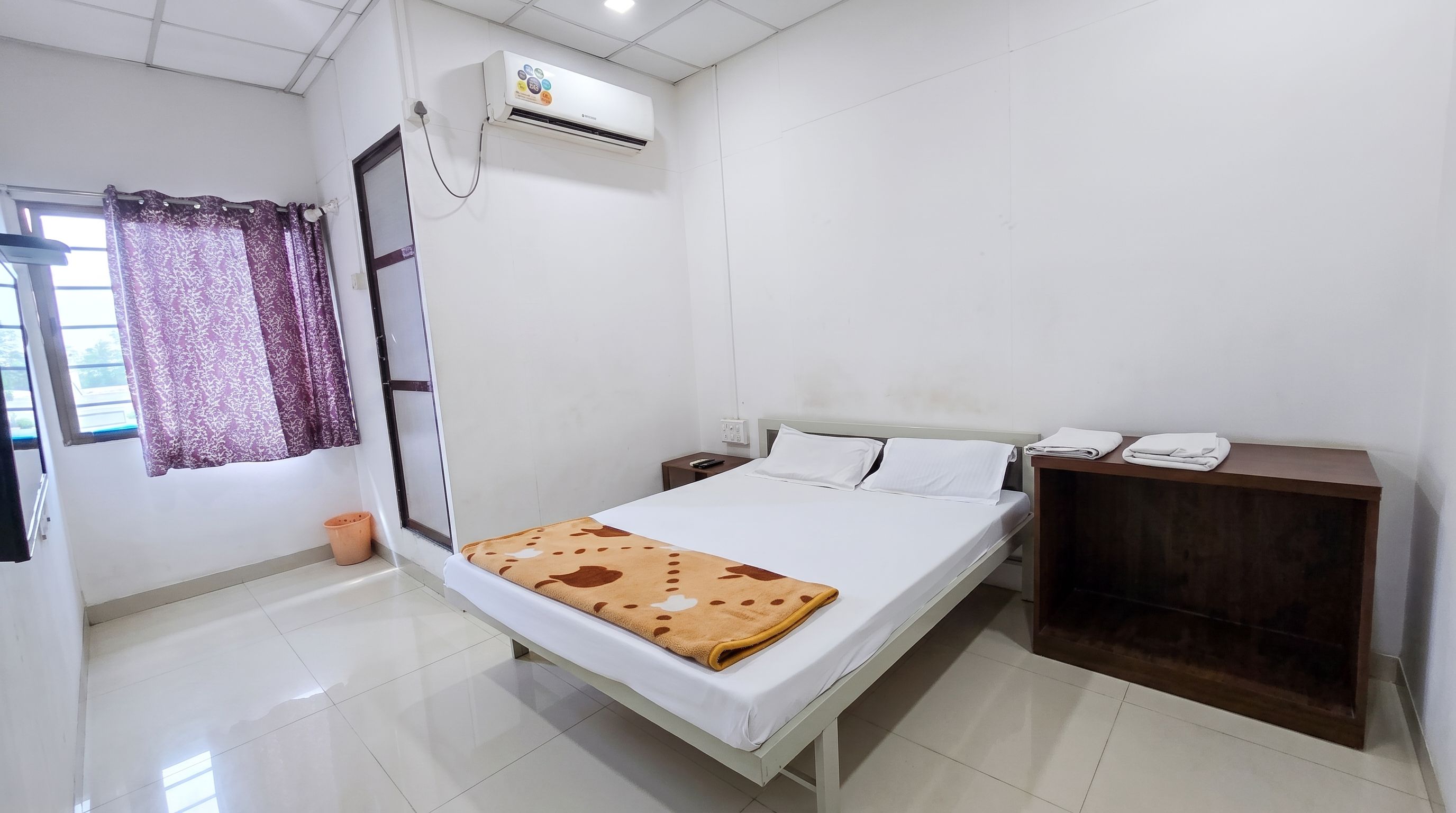 A hotel room with a bed, a nightstand, an air conditioning unit, a window with curtains, and a wooden cabinet.