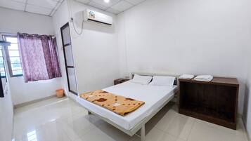 A hotel room with a bed, a nightstand, an air conditioning unit, a window with curtains, and a wooden cabinet.