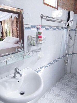 Uma casa de banho com uma pia branca, uma prateleira de vidro com produtos de higiene pessoal, zona de duche e uma janela com vista para um muro de tijolos.