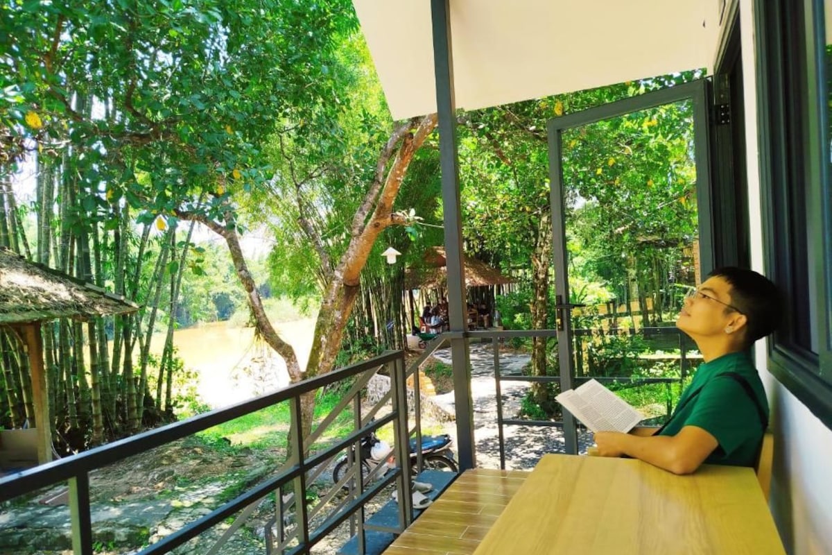 Una persona sentada a una mesa con vista a un paisaje natural, que incluye árboles y un sendero.