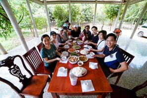 Un grupo de personas sentadas alrededor de una mesa de madera con varios platos, en un espacio al aire libre con techo y árboles al fondo.