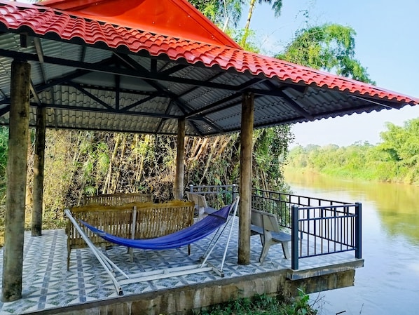 A covered outdoor area with a hammock, a bench, and a railing overlooking a river.