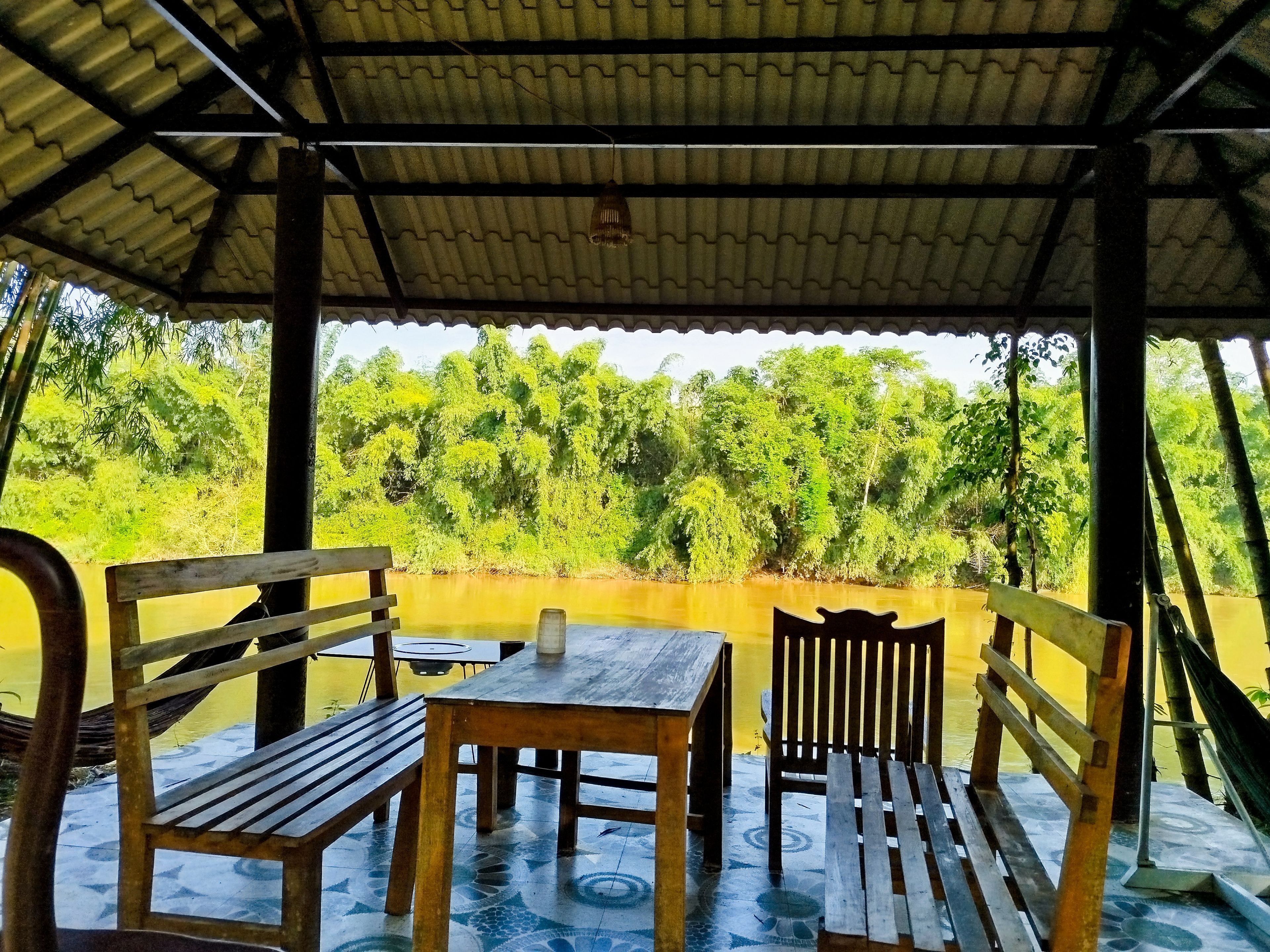 A covered outdoor seating area with wooden furniture and a view of a river and greenery.