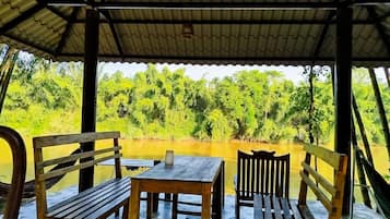 A covered outdoor seating area with wooden furniture and a view of a river and greenery.