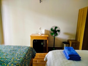 Una habitación de hotel con dos camas, una mesita de noche, un pequeño escritorio con un ventilador y un aplique de pared.