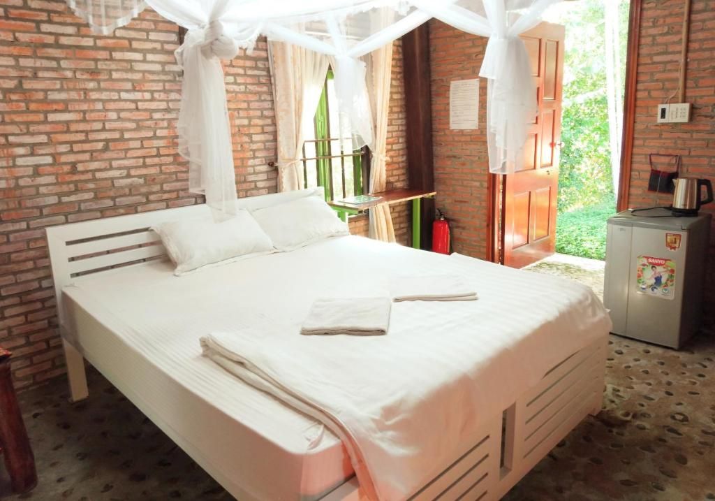 Una cama con dosel, una mesita con una silla, un refrigerador y un muro de ladrillo.