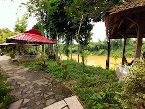 A stone pathway leading to a red-roofed pavilion surrounded by lush greenery and a river.