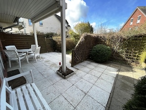 A patio area with a white table and chairs, a small garden bed with a red flower, and a brick wall in the background. - Holiday Apartment Seehund – Arrive. Unwind. Enjoy the Sea. (Schönberg (Holstein))