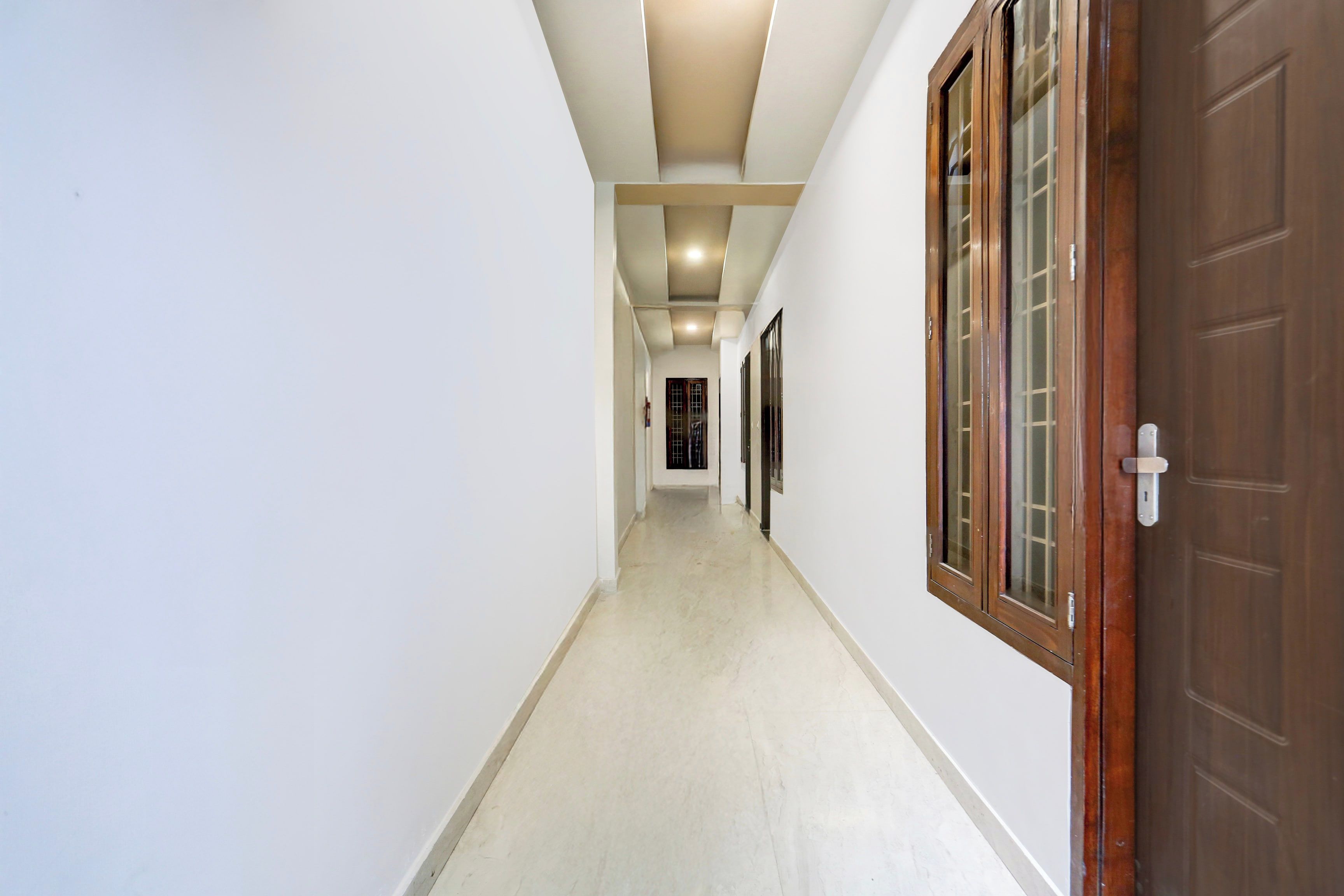 A corridor with a wooden door on the right, white walls, and a tiled floor.