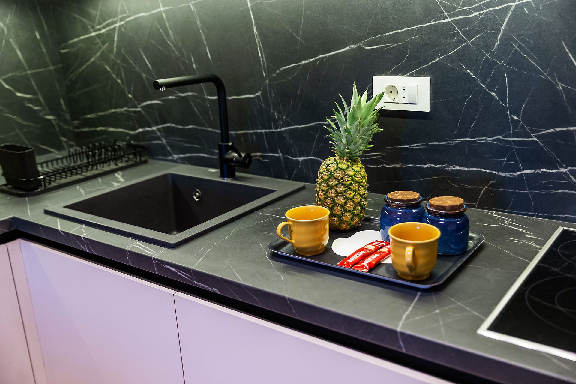 A kitchen counter with a black sink, a pineapple, two mugs, and jars.