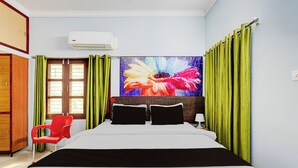 Quarto de hotel com cama, uma cadeira vermelha, um quadro floral grande e cortinas verdes.