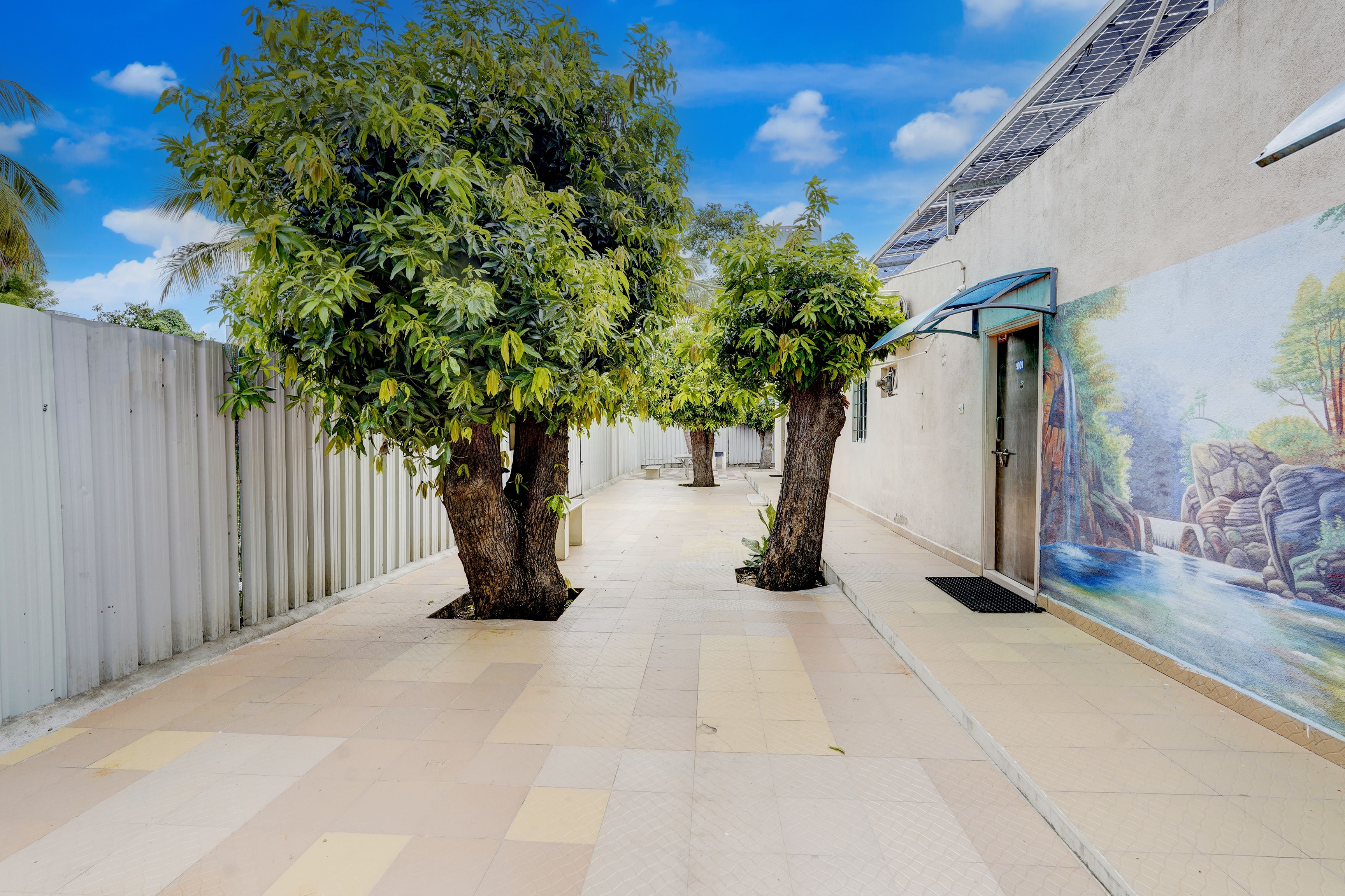 A corridor with tiled flooring, two trees, a white fence, and a mural on the wall.