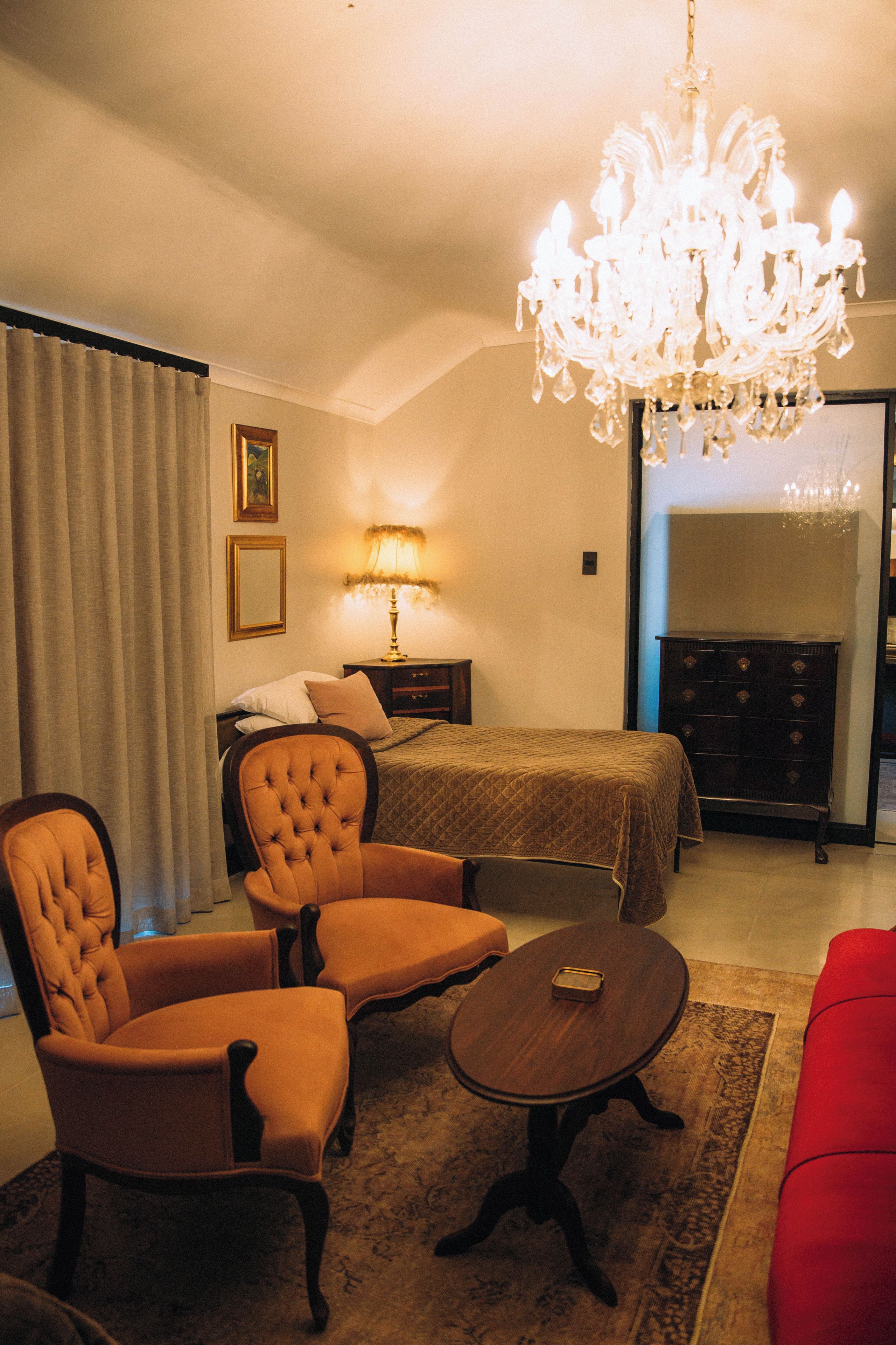 A hotel room with a bed, two armchairs, a red sofa, a chandelier, a lamp, a painting, and a fireplace.