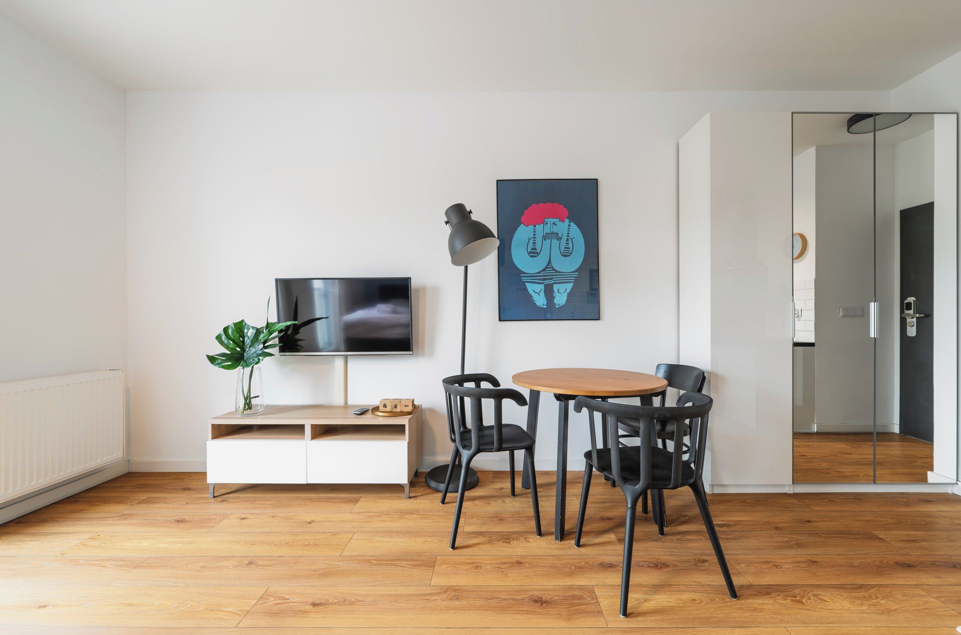 Ein modernes Wohnzimmer mit Fernseher, Couchtisch und vier Stühlen. Auf einem Beistelltisch steht eine Pflanze, an der Wand hängt ein Gemälde, und eine Tür führt in einen anderen Raum.
